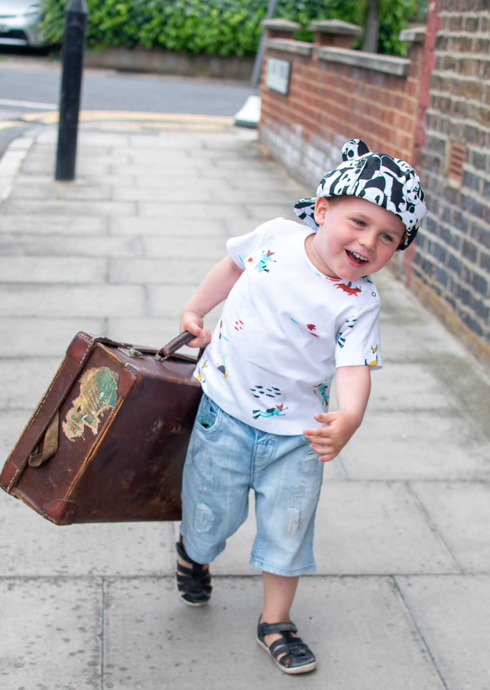 Laurie carrying his suitcase, holiday ready with an outfit from Loopster. He wears an underwater themed T Shirt with jean shorts and a print hat.