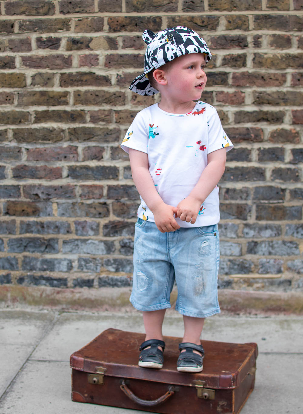 Laurie stood on a suitcase, holiday ready with an outfit from Loopster. He wears an underwater themed T Shirt with jean shorts and a print hat.