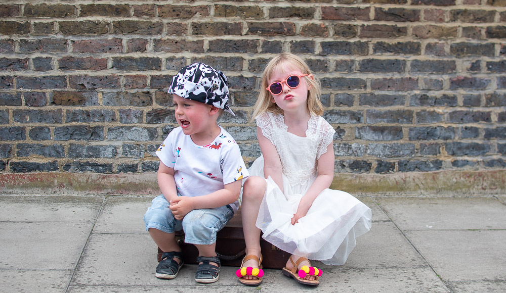 Daisy and Laurie sat on their suitcase holiday ready in their preloved children's clothes from Loopster. Daisy wears a white dress and sunglasses. Laurie an underwater themed T Shirt with jean shorts and a print hat.