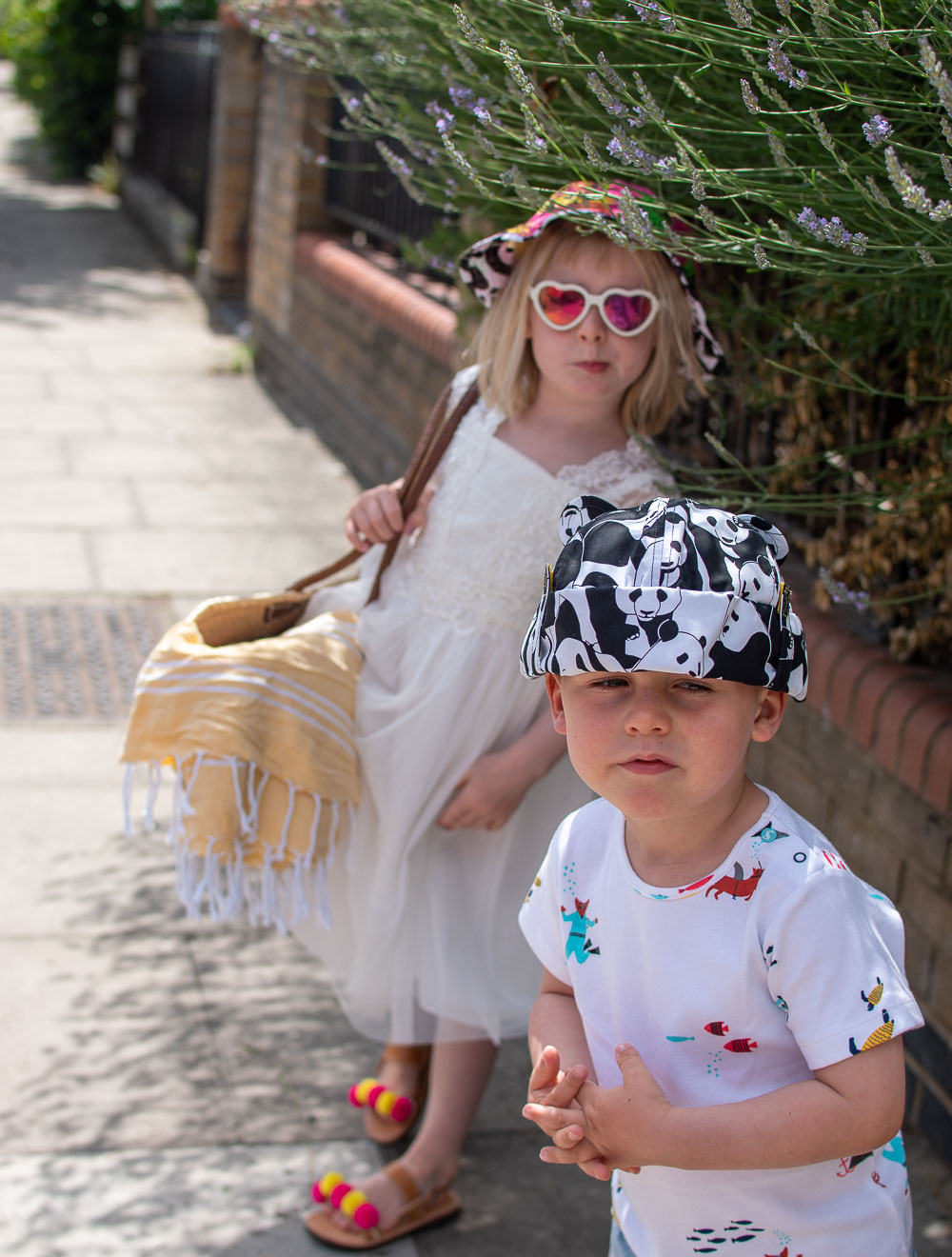 Daisy and Laurie sat on their suitcase holiday ready in their preloved children's clothes from Loopster. Daisy wears a white dress, printed hat and sunglasses. Laurie an underwater themed T Shirt with jean shorts and a print hat.
