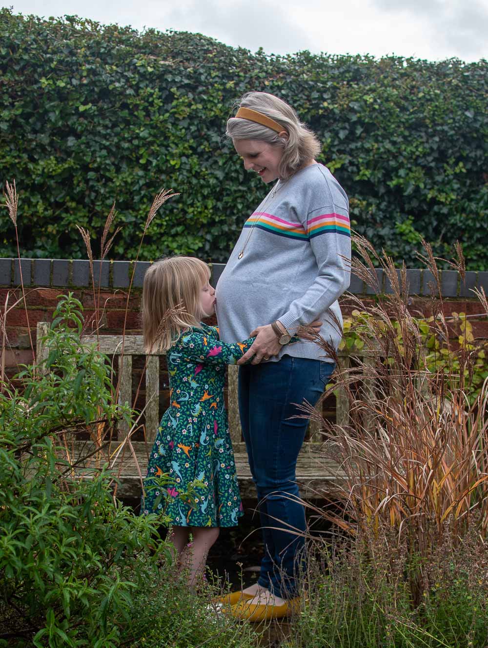 Karen Maurice of n4mummy wearing a rainbow stripped maternity jumper from Frugi and skinny maternity jeans from Jo Jo Mamam Bebe, her daughter Daisy wearing a skater dress from Frugi, kissing her mumimy's bump